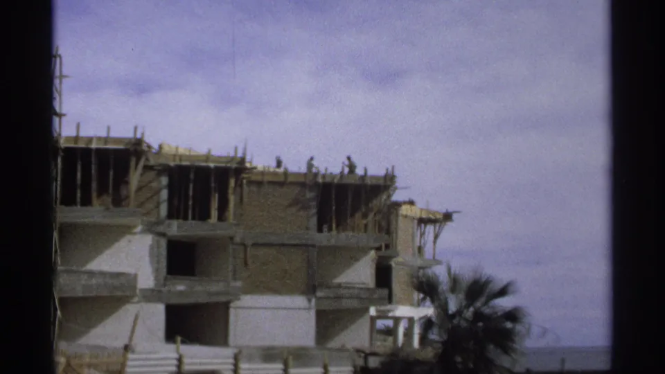 1975 Guaymas, Mexico 8mm frame showing a multi-story building under construction with workers, scaffolding, palm fronds, and ocean beyond.
