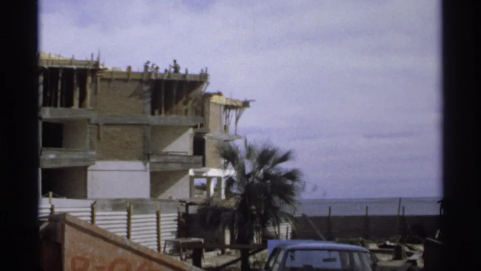 1975 Guaymas 8mm view of a coastal building under construction, with workers above, a palm tree in front, and a parked car near the street.