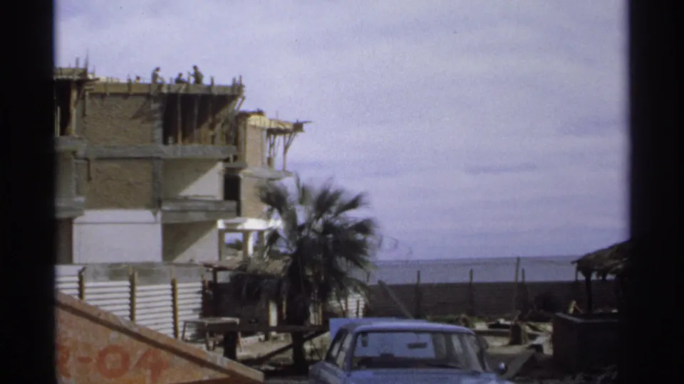 1975 Guaymas home-movie frame showing workers on a partially built concrete structure, with palm tree, parked car, wall, and ocean behind.