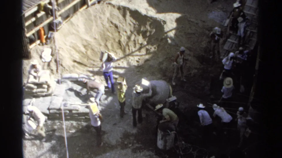Elevated 1975 Guaymas view of workers at a construction site with a cement mixer, stacked bags, and timber scaffolding.