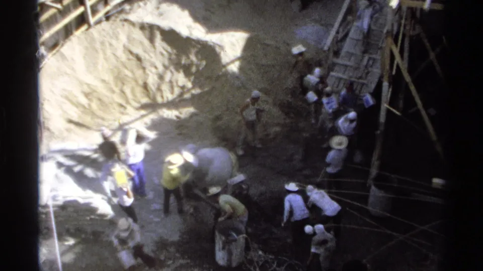 1975 Guaymas construction scene with workers carrying materials near a sand pile, cement mixer, rope lines, and scaffolding.