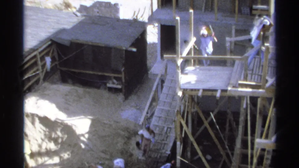 1975 Guaymas construction scene with a small dark shed, timber railings, scaffolding, ladders, and workers in light clothing.