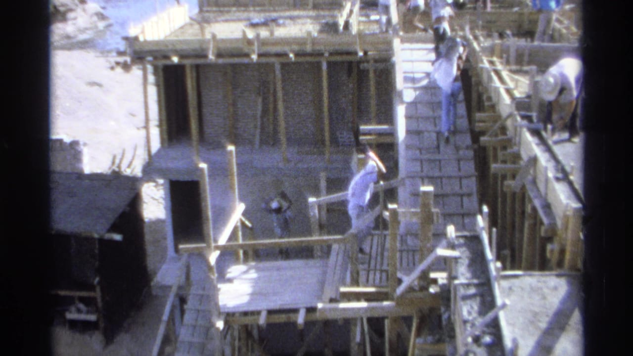Workers move around a seaside construction site with brick walls, timber formwork, and a wooden ramp.