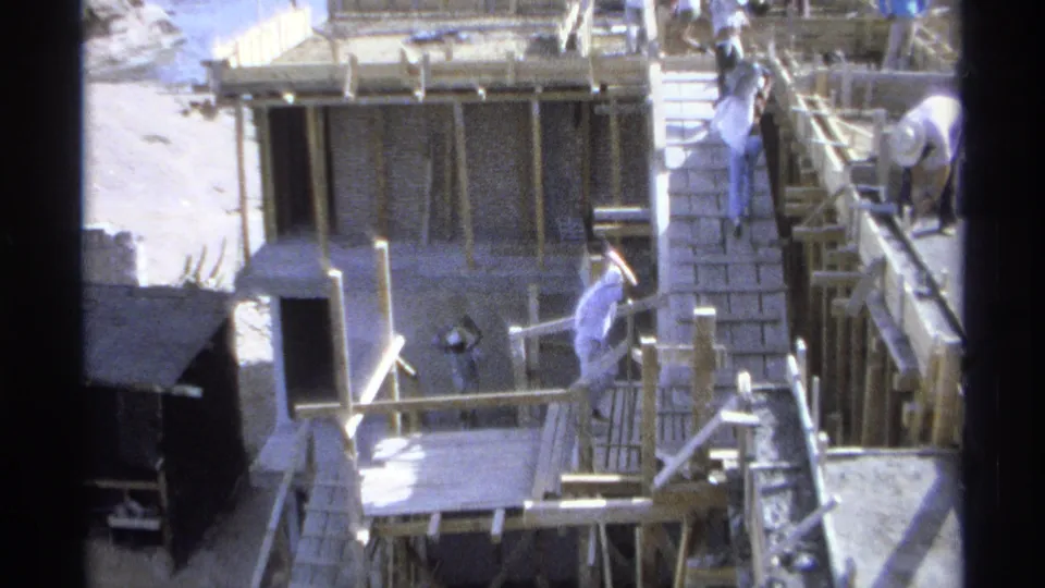 1975 Guaymas, Mexico 8mm frame showing workers at a coastal construction site with exposed brick, timber forms, buckets, and ocean nearby.