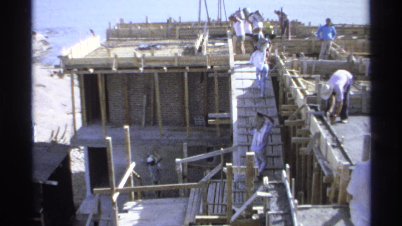 Men work on and around a wooden ramp at an unfinished seaside building site.