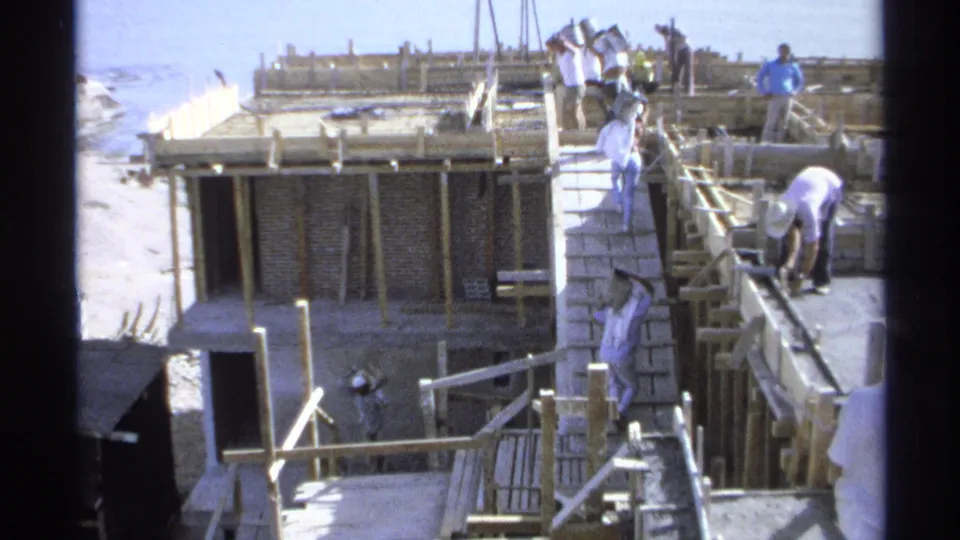 1975 Guaymas, Mexico frame of workers carrying materials across a wooden ramp and formwork near the ocean under bright sun.
