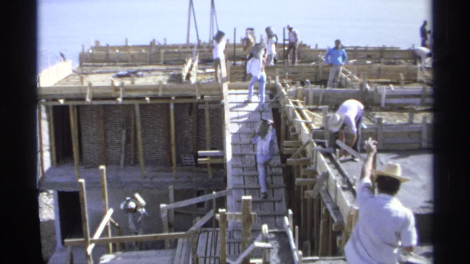 Wider 1975 Guaymas, Mexico 8mm view of a beachside construction site with workers in white clothing, masonry, formwork, and sea behind.