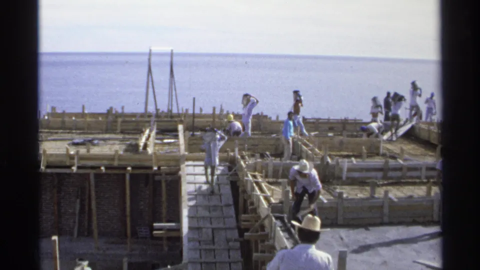 1975 Guaymas, Mexico frame showing workers on timber formwork and a ramp, with bright ocean water behind the unfinished structure.