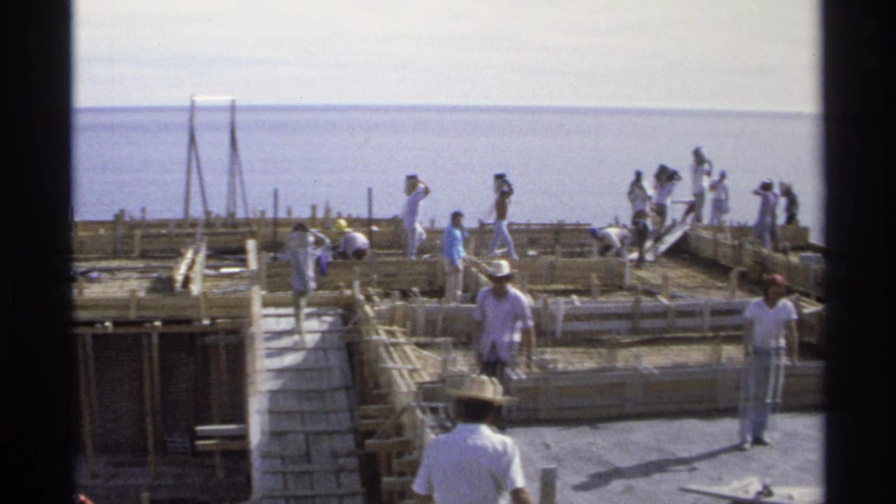 Workers in hats and light clothing build wooden forms for a concrete structure by the ocean in Guaymas, Mexico, 1975.