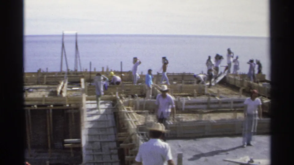 Workers in hats and light clothing build wooden forms for a concrete structure by the ocean in Guaymas, Mexico, 1975.