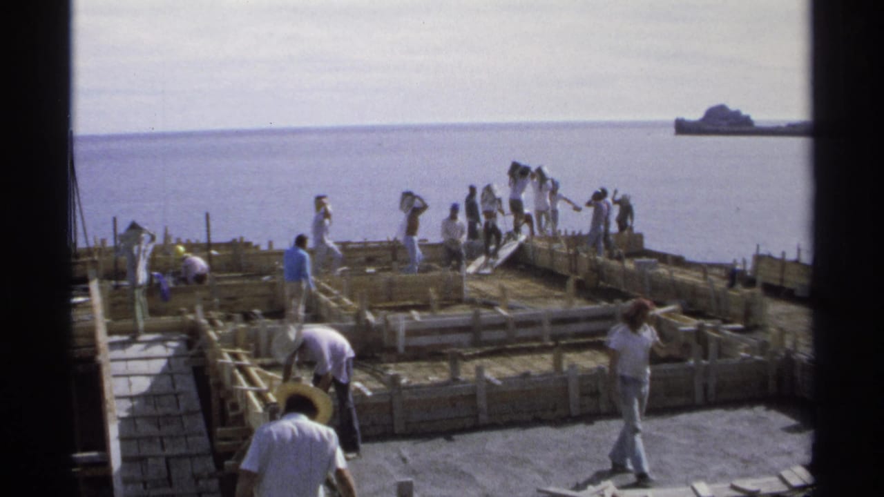 Workers continue construction around low wooden forms with the ocean in the background.