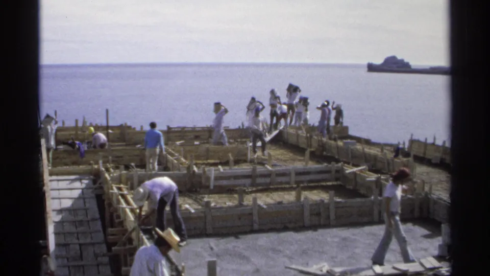 Workers are spread across a coastal construction site in Guaymas, Mexico, with wooden forms and the sea visible behind them.