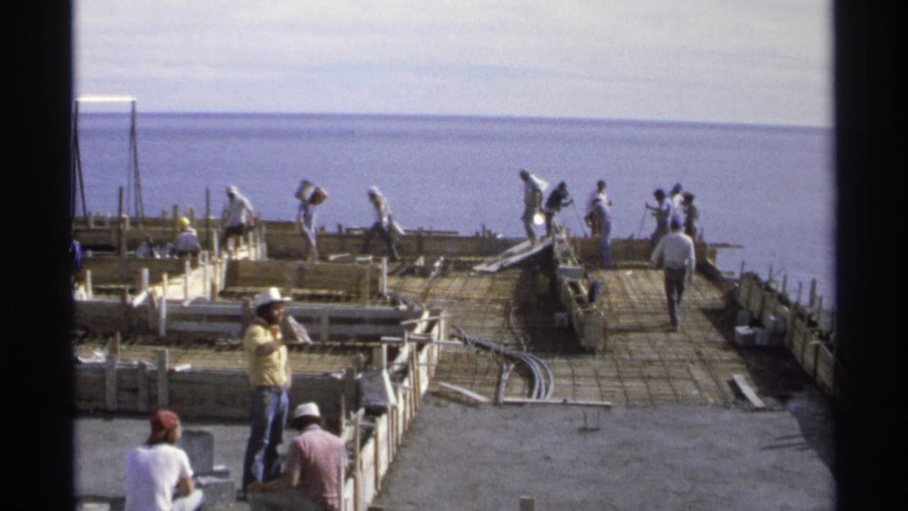 Workers on a rebar foundation near the ocean, with hard hats, formwork, and dark film borders visible.