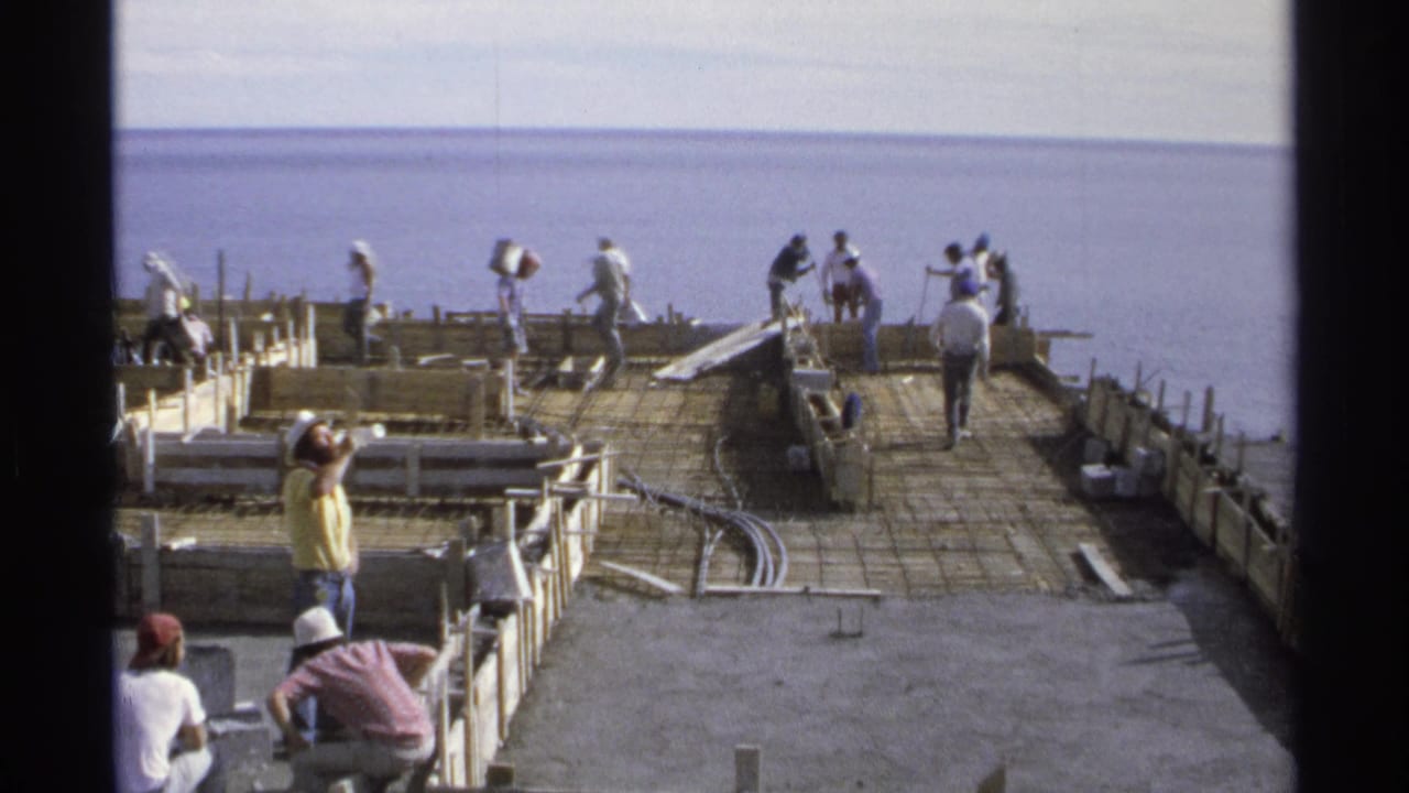 Construction crew working across a large foundation beside the ocean, with equipment and materials on the slab.