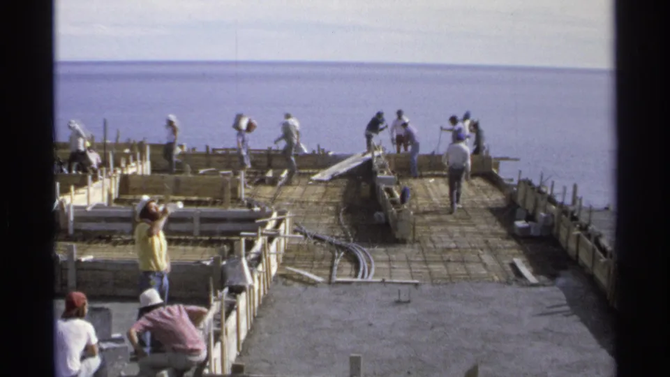 1975 Guaymas construction scene with workers spread across a rebar grid near the water, viewed through an 8mm contact-frame border.