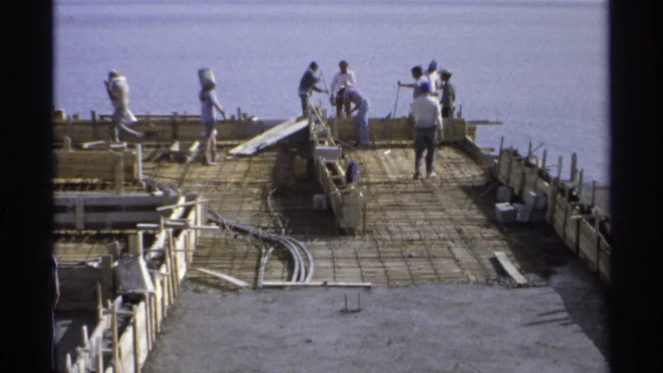 1975 Guaymas, Mexico frame showing workers on a coastal foundation, including hard hats, rebar, formwork, and scattered building materials.