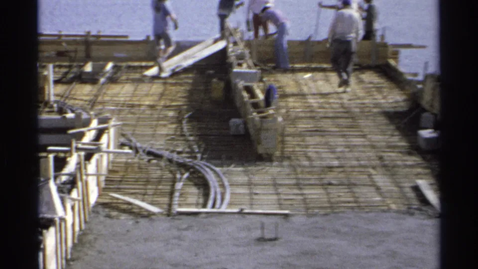 1975 Guaymas construction frame showing a closer view of rebar, wood forms, workers, and foreground hoses or cables near the ocean.