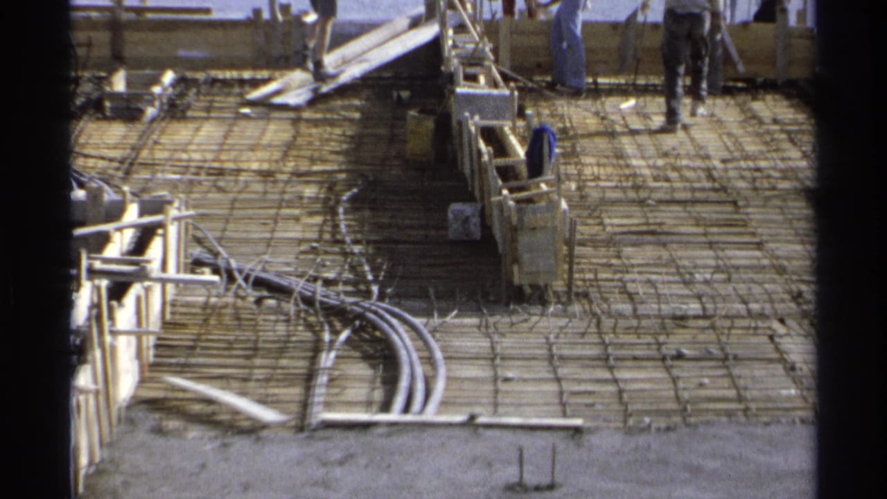 Rebar grid and wooden forms on a sunlit construction foundation with workers partly visible at the edge.