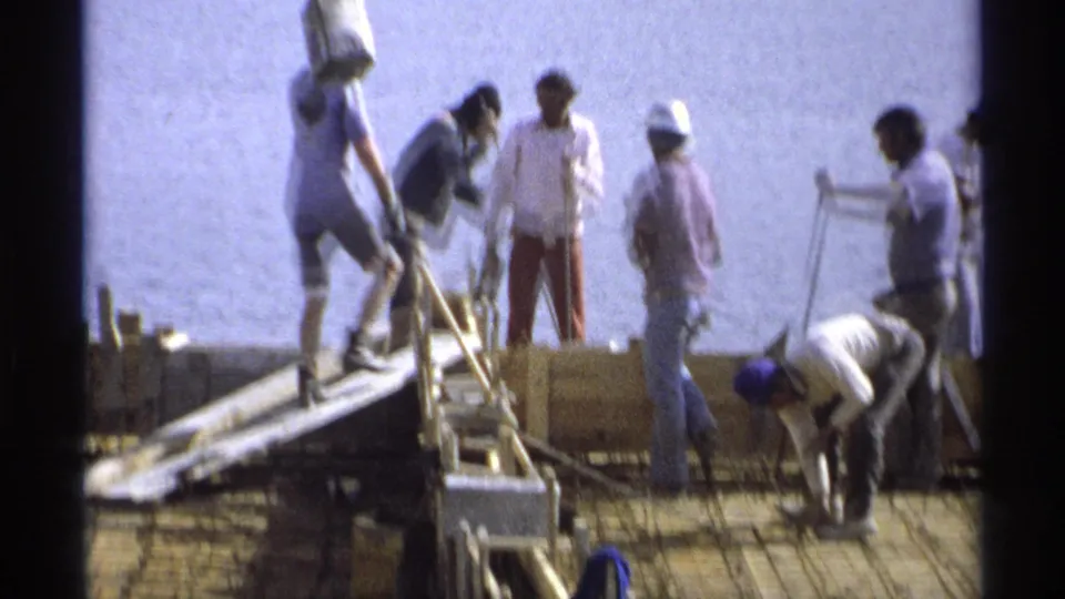 1975 Guaymas footage shows construction workers in hard hats and casual clothes handling forms near a ladder and rebar deck.