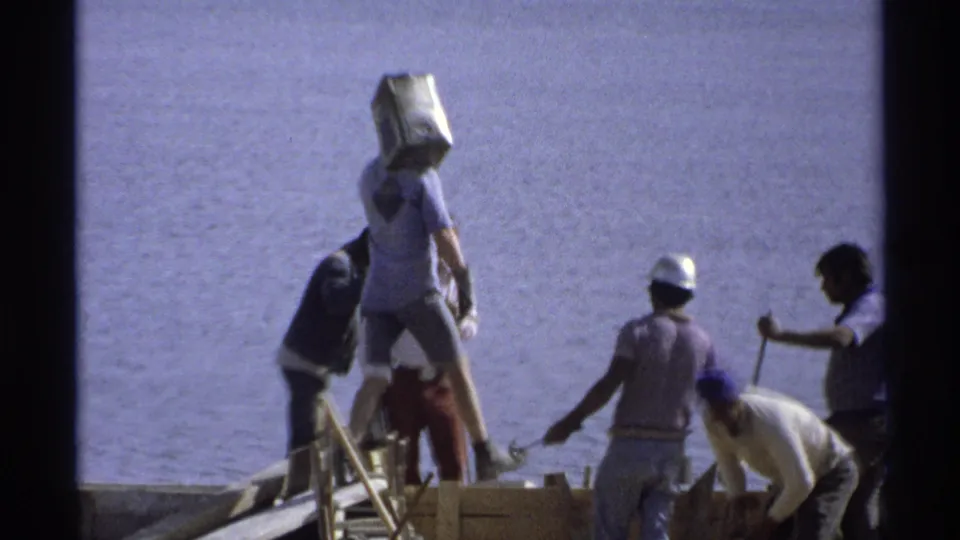 At the Guaymas site in 1975, a worker carries a metal bucket as others bend and stand near wooden foundation forms.