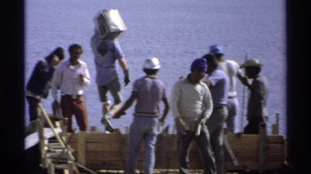 1975 Guaymas construction workers on a waterfront foundation, with hard hats, tools, wood forms, and water in the background.