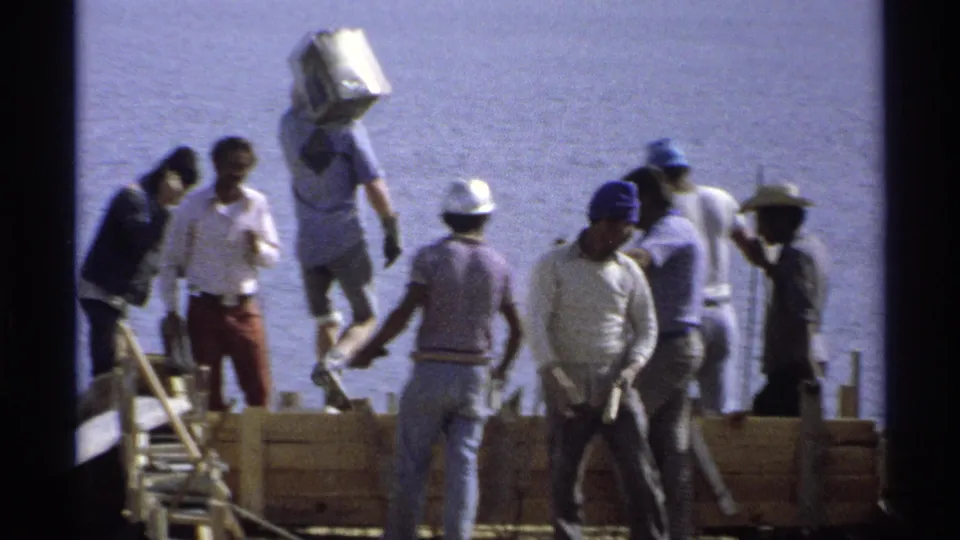 1975 Guaymas construction workers on a waterfront foundation, with hard hats, tools, wood forms, and water in the background.