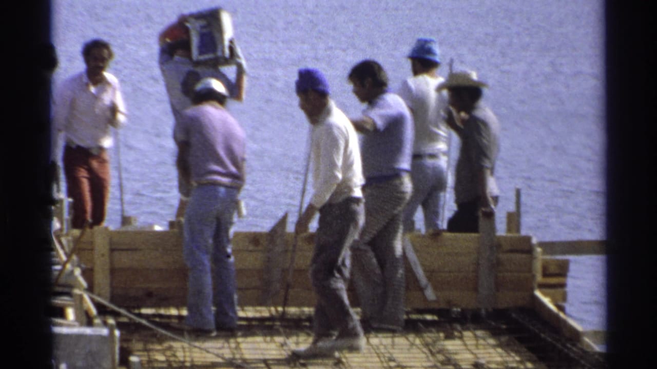 Several workers stand and move along a wooden foundation form with water behind them.