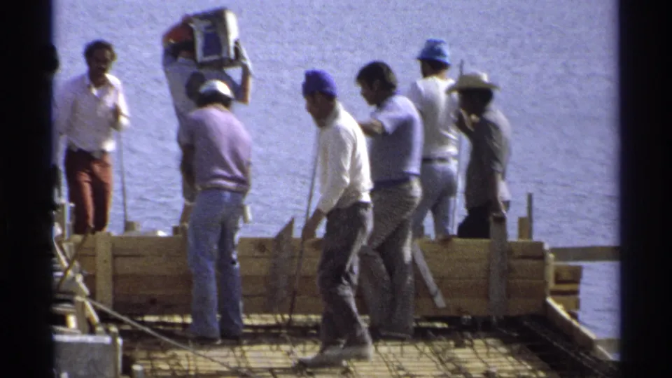 Workers in casual clothing and hard hats handle tools and materials on a waterfront construction form in Guaymas, Mexico.