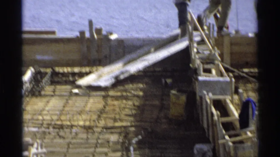 Close view of the construction platform showing rebar, wooden forms, ladders, and partial worker figures beside the waterfront.