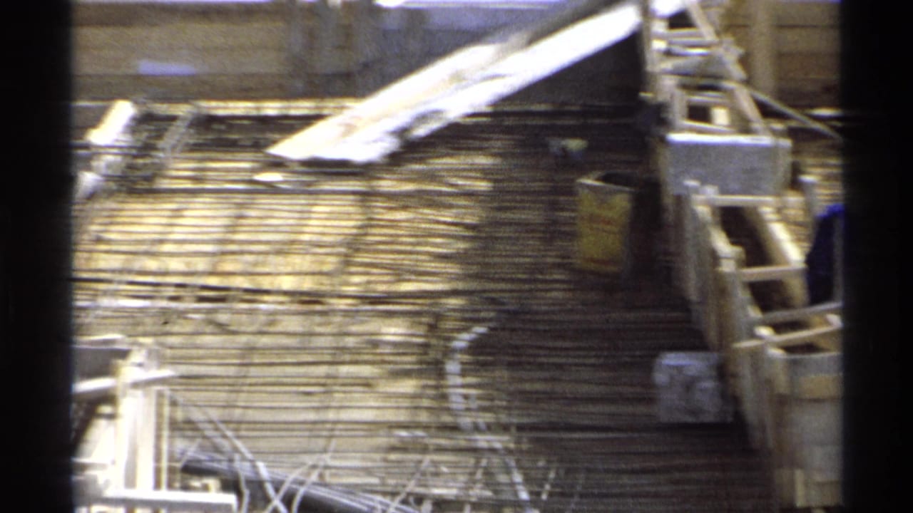 1975 Guaymas construction site seen from above with rebar grids, timber formwork, a ladder, and partial workers at the frame edges.