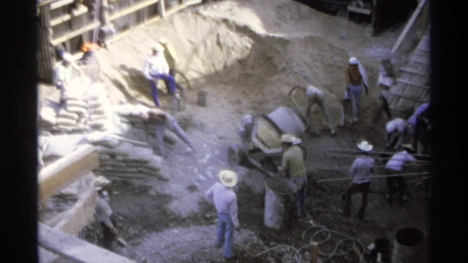 Several workers move around a Guaymas construction pit with sand piles, shovels, buckets, timber supports, and a small cement mixer.