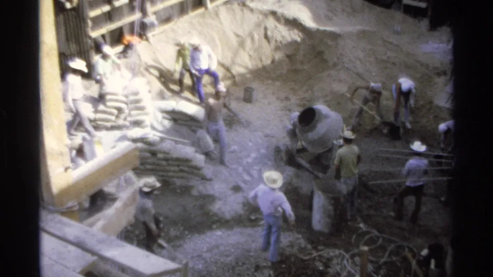 Workers in hats and casual clothing handle materials around a cement mixer, sand piles, timber supports, and rebar in 1975 Guaymas.