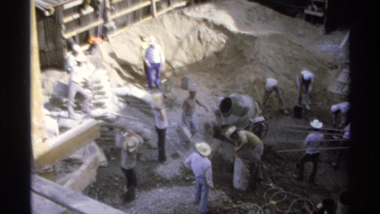Men shovel near a cement mixer beside a large mound of sand or gravel.
