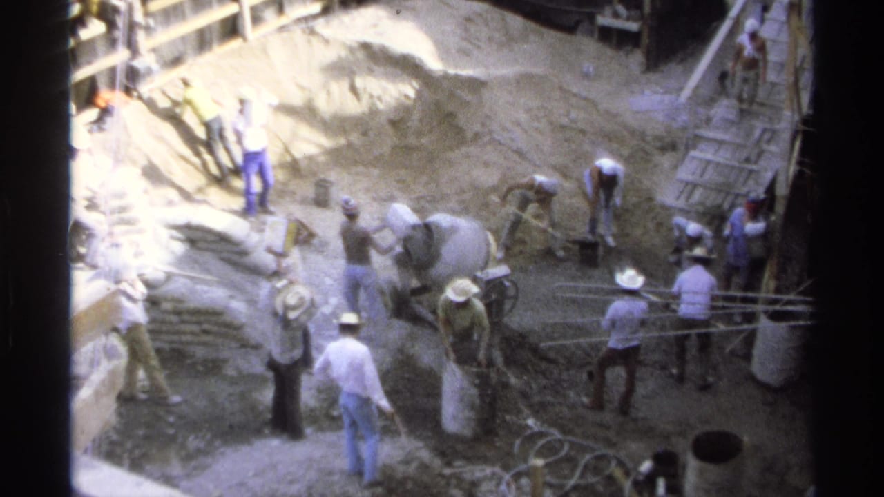 Workers shovel and handle materials beside a cement mixer and excavation area.