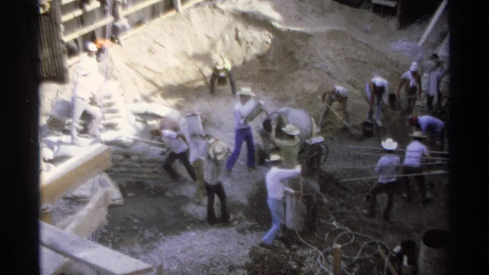 1975 Guaymas building-site activity with workers around a cement mixer, sand mound, and hand tools in bright sun.