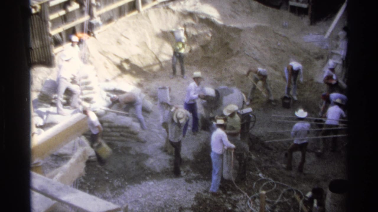 Workers in light clothing and hats handle materials at a 1975 Guaymas construction site seen from above.