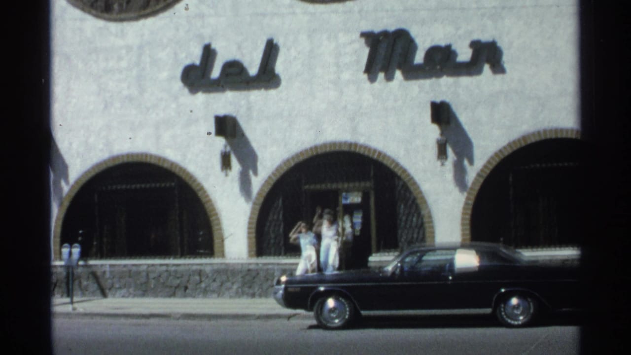 Pedestrians wave near arched entrances of a white building while a black car is parked at the curb.