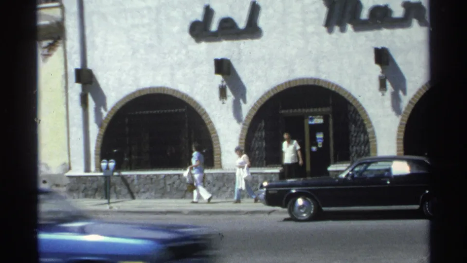 1975 Guaymas sidewalk view with pedestrians passing a white arched facade, parking meters, and a black car near the curb.