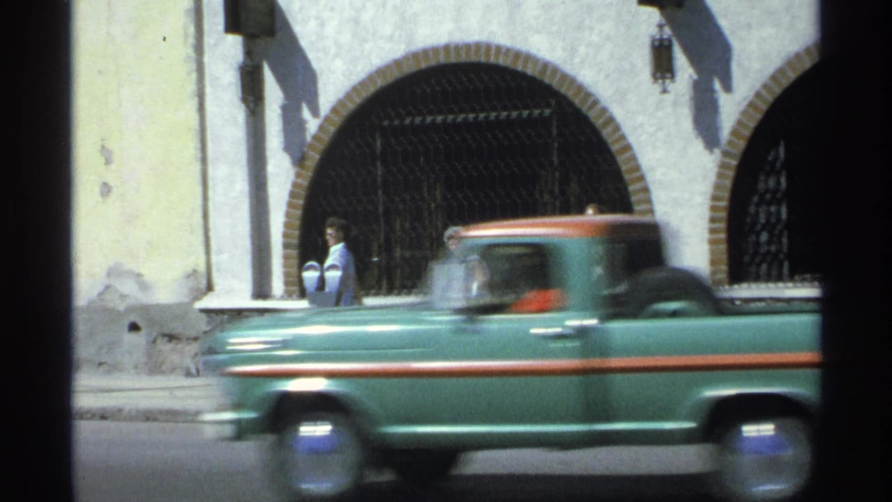In Guaymas, Mexico, a green vintage pickup moves past a yellow stucco facade with arched openings and metal gates.