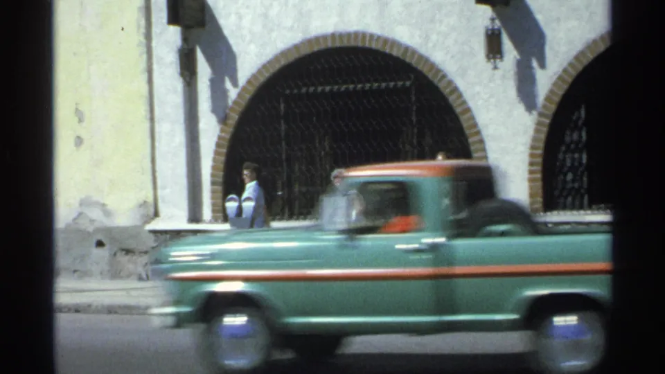 In Guaymas, Mexico, a green vintage pickup moves past a yellow stucco facade with arched openings and metal gates.