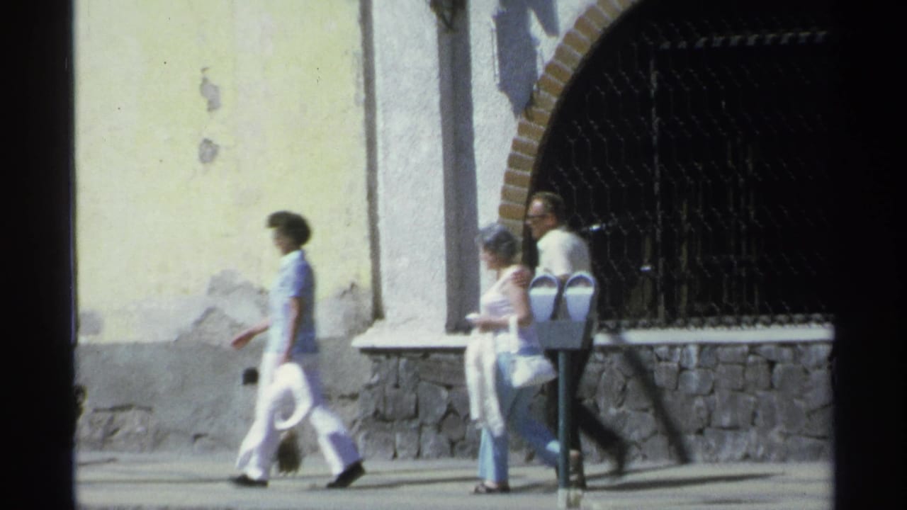 Three adults walking along a sidewalk beside a yellow wall and arched gate.