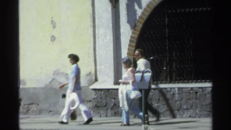 Three adults walk along a Guaymas sidewalk in 1975 beside a yellow wall, stone base, arched opening, and dark metal gate.