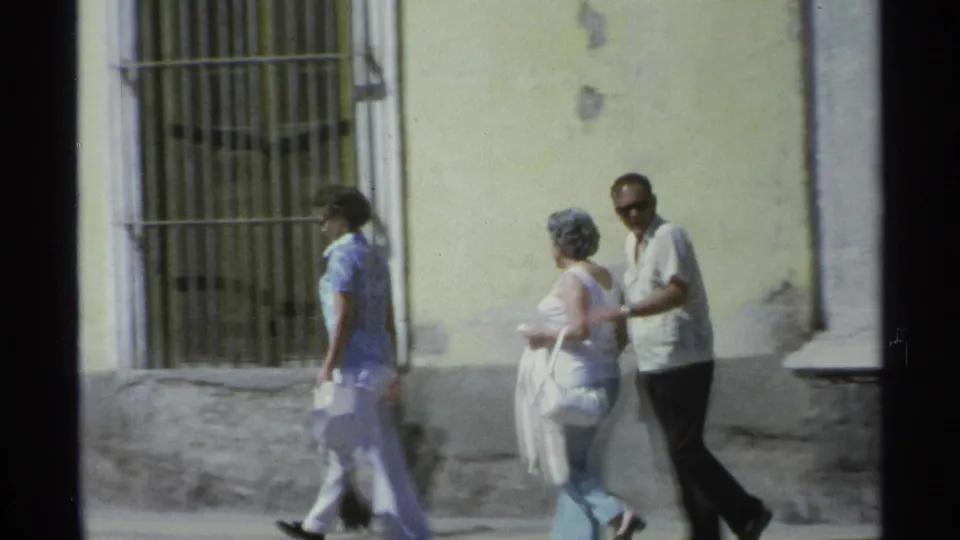 Two women and a man continue along the Guaymas sidewalk, passing a barred window set into a weathered yellow facade.