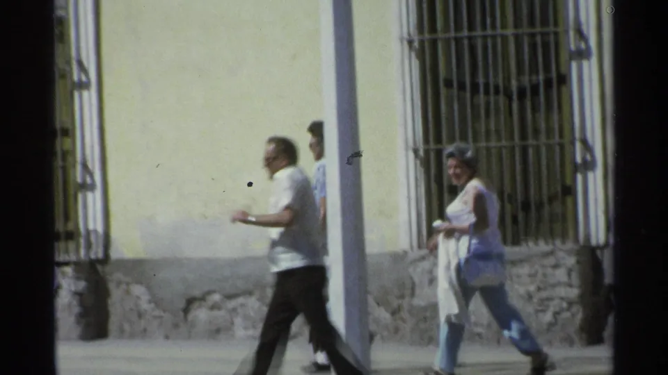 A man and woman walk through the frame in Guaymas, Mexico, with a barred window, utility pole, and weathered wall behind them.