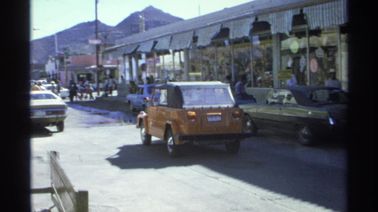 1975 Guaymas street scene with an orange Volkswagen Thing, other period cars, pedestrians, storefront awnings, and mountains beyond.