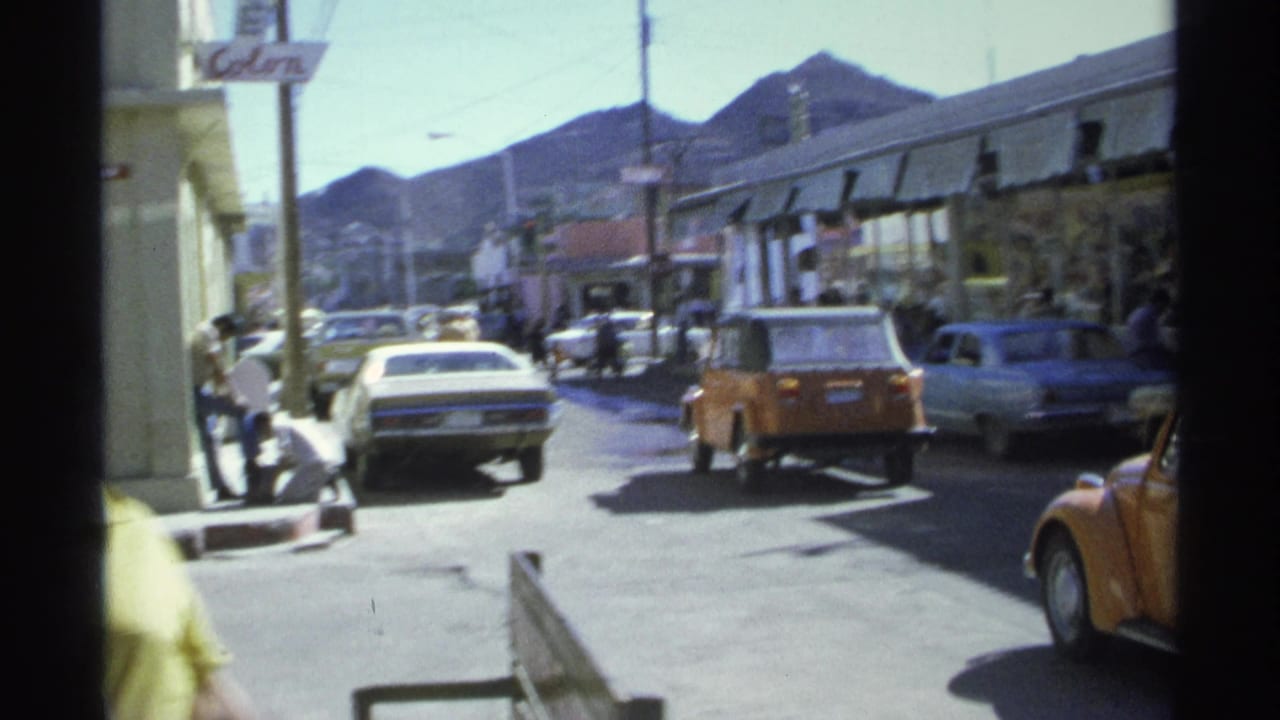 Street traffic passes storefronts as pedestrians walk near a curb with mountains in the distance.