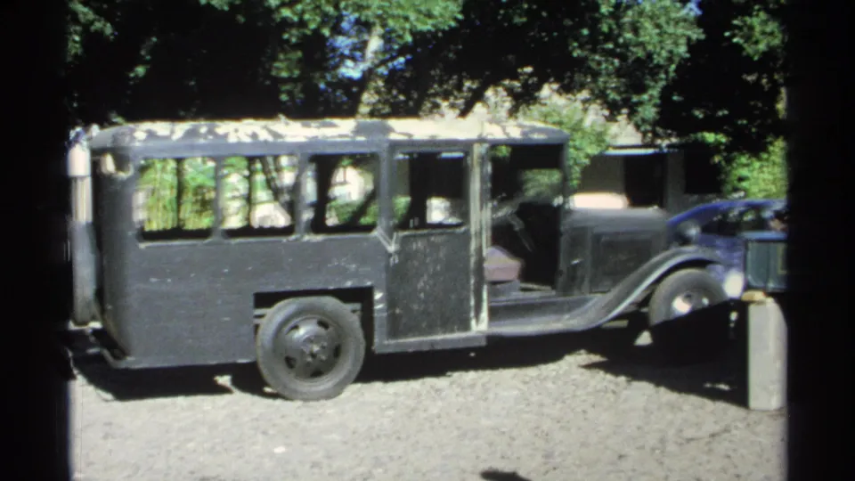 A black vintage bus sits on a gravel lot in the 1975 Guaymas footage, with trees, shade, and part of another vehicle nearby.