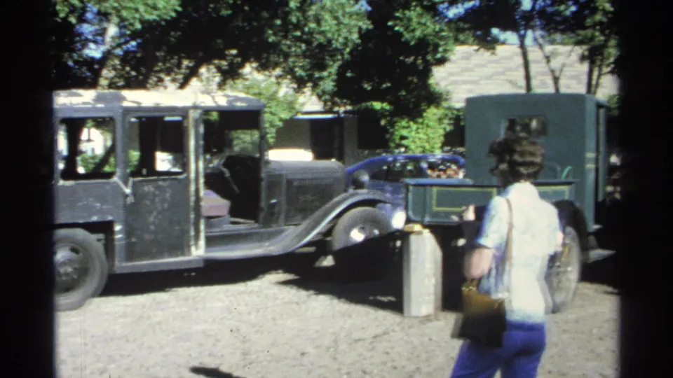 In the 1975 Guaymas home-movie footage, a woman in period casual clothing looks toward a black vintage bus on a gravel lot.
