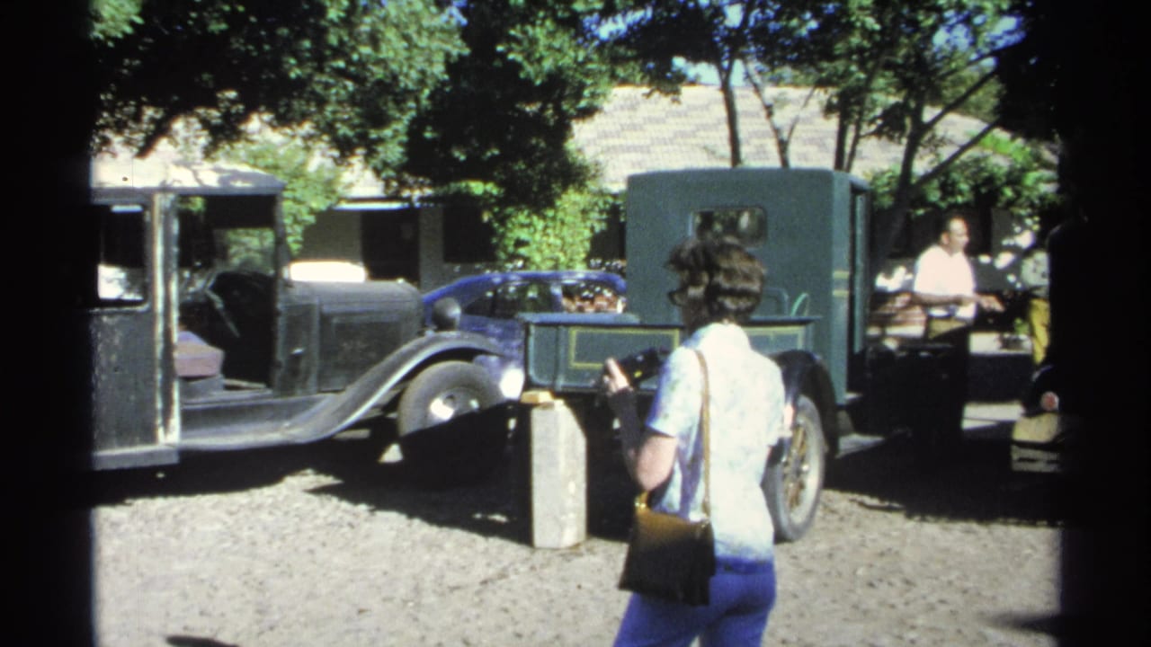 In Guaymas, Mexico, a woman walks through a shaded gravel courtyard past older automobiles and adults standing nearby.