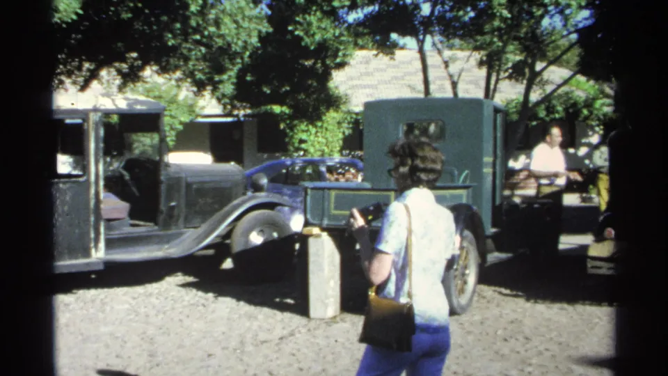 In Guaymas, Mexico, a woman walks through a shaded gravel courtyard past older automobiles and adults standing nearby.